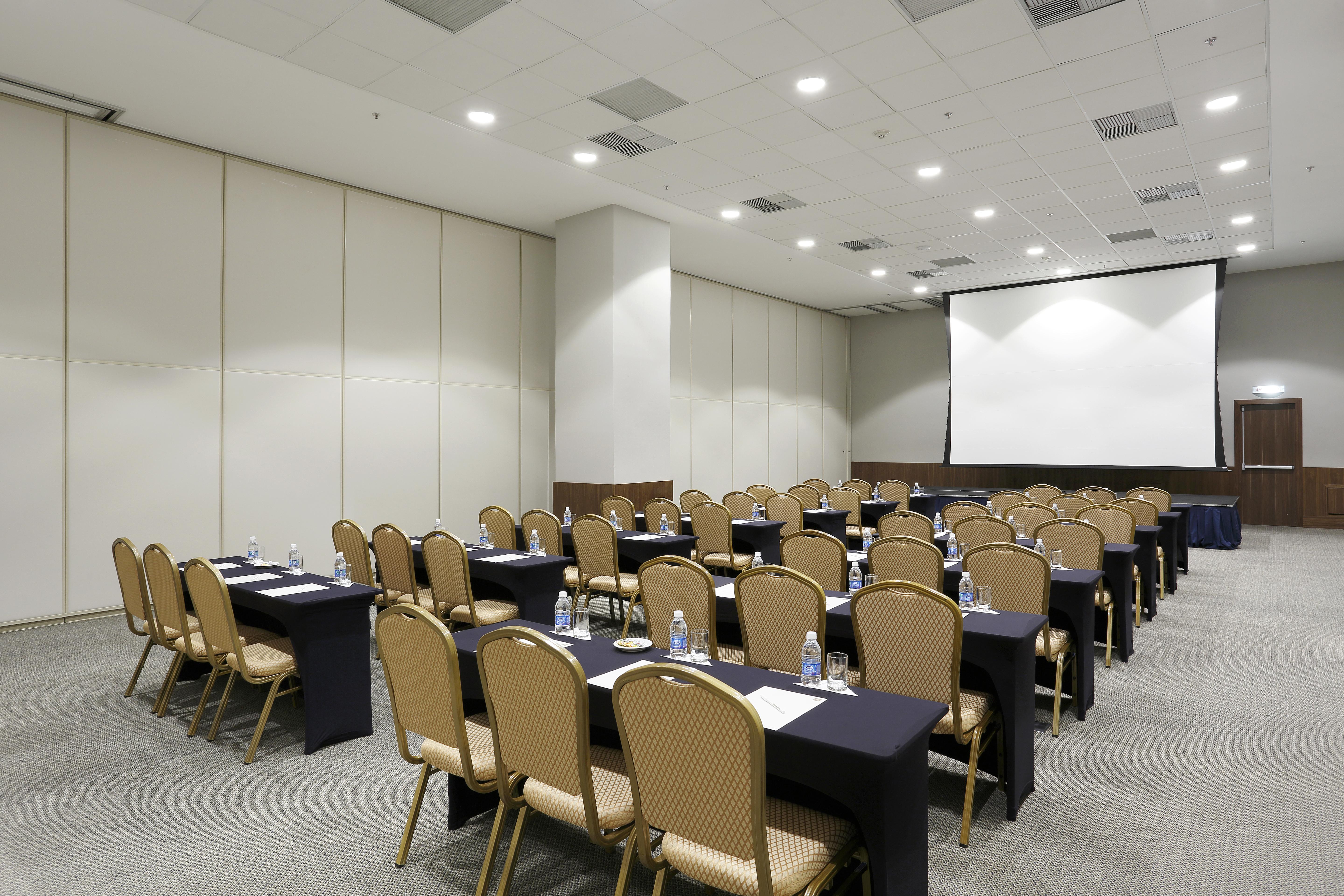 Windsor Marapendi Hotel Rio de Janeiro Zewnętrze zdjęcie The photo shows a conference or meeting room arranged with several rows of tables and chairs. The tables are covered with dark tablecloths and have water bottles placed on them. The room is bright and spacious, featuring a large projection screen on 